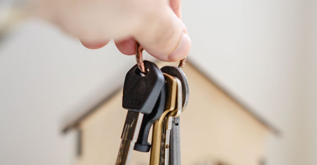 Close-up of a hand holding keys with a miniature wooden house in the background, symbolizing real estate investment.